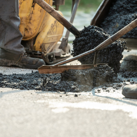 Two construction workers working together to patch a bump in the road with fresh asphalt.